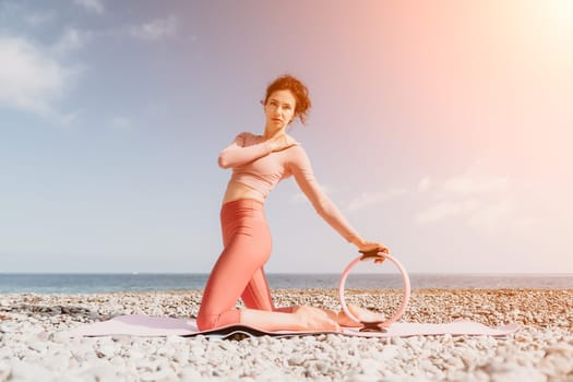 Middle aged well looking woman with black hair doing Pilates with the ring on the yoga mat near the sea on the pebble beach. Female fitness yoga concept. Healthy lifestyle, harmony and meditation.