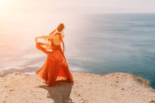 Side view a Young beautiful sensual woman in a red long dress posing on a rock high above the sea during sunrise. Girl on the nature on blue sky background. Fashion photo.