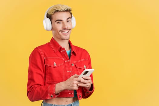 Gay man looking at camera listening to music with mobile in studio with yellow background