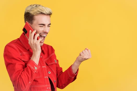 Gay man celebrating while talking to the mobile in studio with yellow background