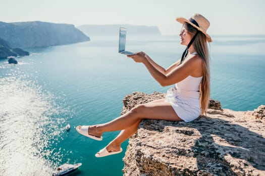 Working remotely on seashore. Happy successful woman female freelancer in straw hat working on laptop by the sea at sunset. Freelance, remote work on vacation