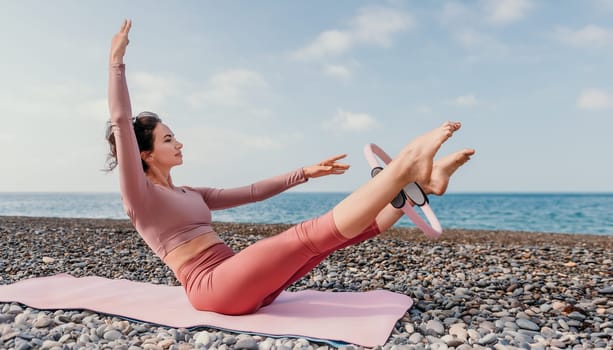 Middle aged well looking woman with black hair doing Pilates with the ring on the yoga mat near the sea on the pebble beach. Female fitness yoga concept. Healthy lifestyle, harmony and meditation.