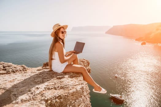 Successful business woman in yellow hat working on laptop by the sea. Pretty lady typing on computer at summer day outdoors. Freelance, travel and holidays concept.