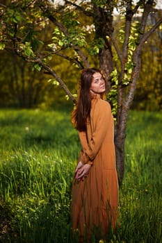 a sweet, attractive woman with long red hair stands in the countryside near a flowering tree in a long orange dress and looks at the camera over her shoulder, with her hands folded on her back. Vertical photography in nature. High quality photo