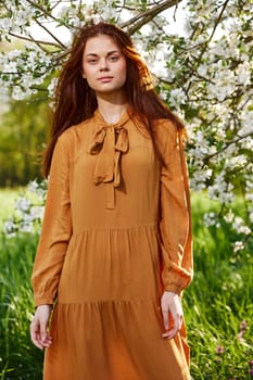 a bright vertical photo of an attractive woman in a long, stylish orange dress standing next to a flowering tree in sunny, warm weather, looking pleasantly into the camera, illuminated from the side. High quality photo