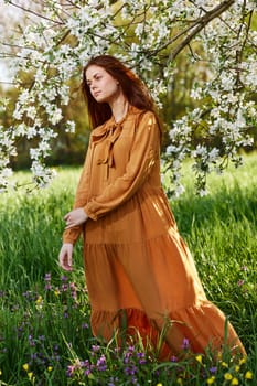 a bright vertical photo of an attractive woman in a long orange dress standing next to a flowering tree in sunny, warm weather. High quality photo