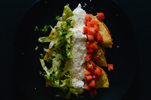 Mashed potato and chorizo fried tacos dorados with lettuce, tomatoes, sour cream and cotija cheese. Shot on black background.