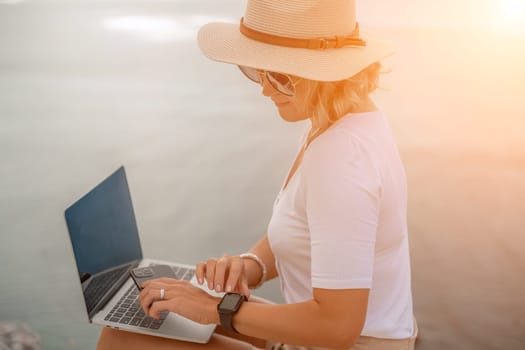 Freelance women sea working on the computer. Good looking middle aged woman typing on a laptop keyboard outdoors with a beautiful sea view. The concept of remote work