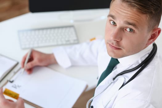 Male medicine doctor hand hold jar of pills and write prescription to patient at worktable. Panacea and life save prescribing treatment legal drug store concept. Empty form ready to be used