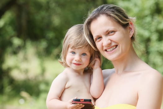 Happy little smiling child in the arms of the mother on a natural background. Holds a smartphone in the hands of watching developing video plays games