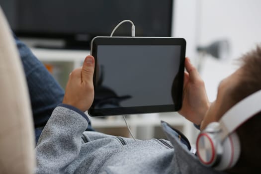 Male hand holds tablet pad in home setting while sitting on the couch engaged an internet surfing using application to press a finger on the display leisure listering music concept closeup.