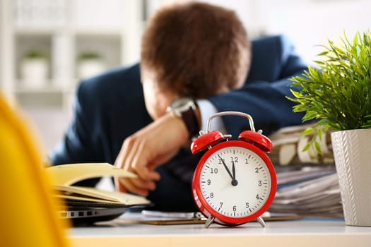 Red alarm clock shows late time closeup and tired office male clerk in suit take nap on table workplace full of exam papers. Career frustration freelance employment fail study problem low energy down