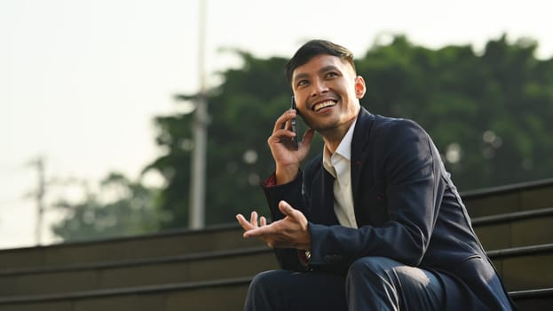 Millennial businessman talking on mobile phone while sitting on stairs in front of office building.
