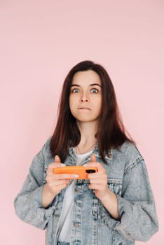 Portrait of a young woman in casual denim jacket with smartphone playing game, isolated on pink background conveying hesitancy and insecurity.