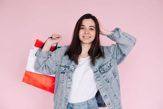 Excited young woman in casual denim shirt raises her arm in celebration, standing in front of a pink background, happy after shopping or finding a great deal online.