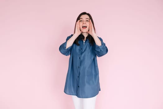 Portrait of young woman with joyous expression, hands near open mouth as if sharing news or telling a funny story, beaming a big toothy smile, isolated on bright pink background