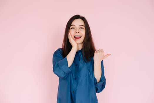 Young woman expressing amazement and joy, touching cheek and pointing finger at empty space, with a big toothy smile on pink isolated background.