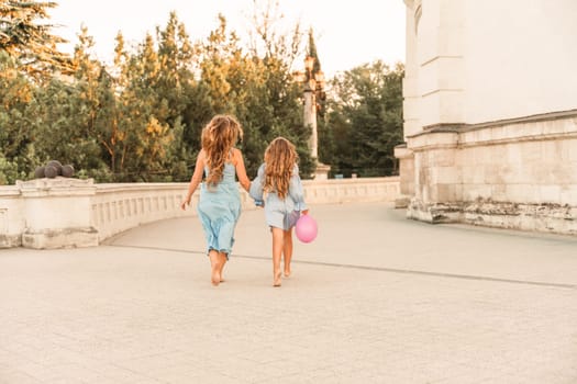 Daughter mother run holding hands. In blue dresses with flowing long hair against the backdrop of a sunset and a white building