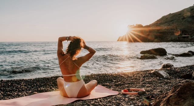 Young woman in swimsuit with long hair practicing stretching outdoors on yoga mat by the sea on a sunny day. Women's yoga fitness pilates routine. Healthy lifestyle, harmony and meditation concept.