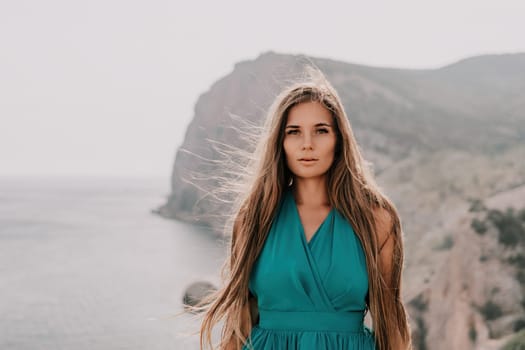 Side view a Young beautiful sensual woman in a mint long dress posing on a volcanic rock high above the sea during sunset. Girl on the nature on overcast sky background. Fashion photo