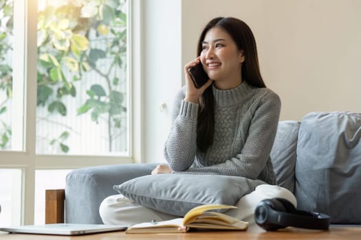 Pretty young asian woman having phone conversation, sitting on couch in living room and using laptop, Cheerful lady resting at home, websurfing and talking on mobile phone.