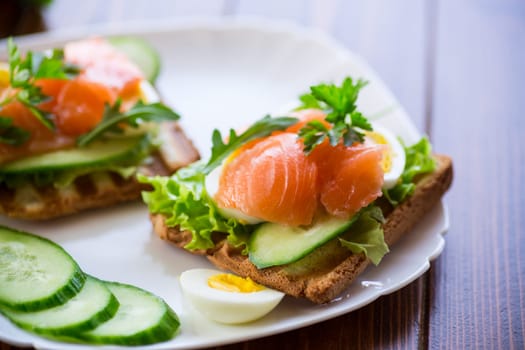 fried toast with lettuce, egg, cucumbers and red fish in a plate, on a wooden table.