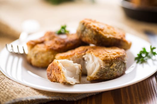 A piece of hake fish fried in a crispy crust, in a plate on a wooden table.