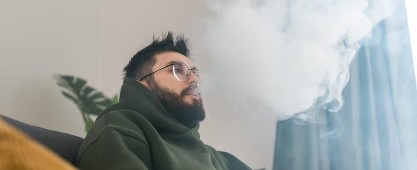Bearded man is smoking hookah at home and blowing cloud of smoke - chill time and resting