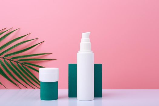 Creative trendy set with two white cosmetic jars with skin care products on green geometric shapes for product presentation against pink background with palm leaf. 