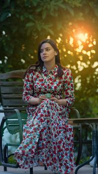 Beautiful Latin woman in a long dress with floral design sitting in the garden with a cup of coffee in her hands during the afternoon with the sunlight passing through the trees in the background out of focus