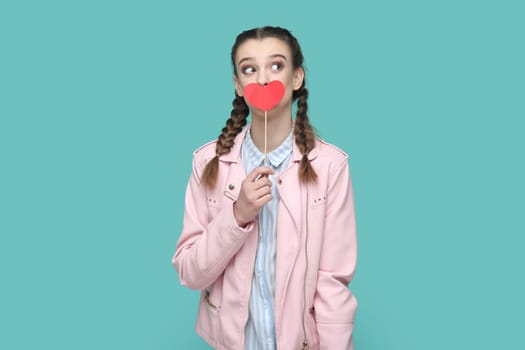 Portrait of funny positive teenager girl with braids wearing pink jacket covering her mouth with red heart on stick, looking away, dreaming. Indoor studio shot isolated on green background.