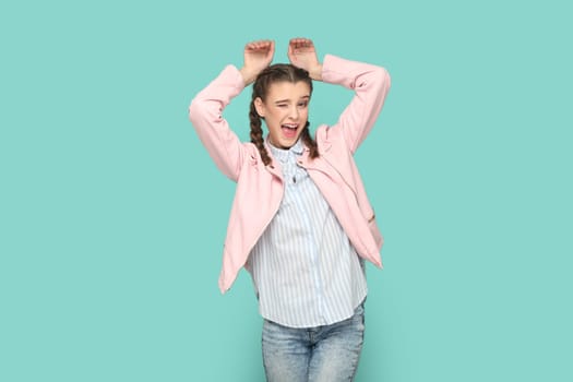 Portrait of funny positive childish teenager girl with braids wearing pink jacket showing bunny ears, winking to camera. Indoor studio shot isolated on green background.