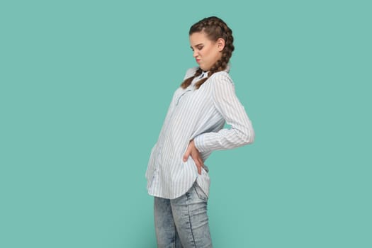 Portrait of dissatisfied teenager girl with braids wearing striped shirt smirks face, keeps hand on back, suffers from terrible pain. Indoor studio shot isolated on green background.