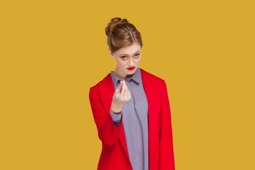 Portrait of serious young adult woman with red lips standing and showing money gesture, asking to give her salary, wearing red jacket. Indoor studio shot isolated on yellow background.