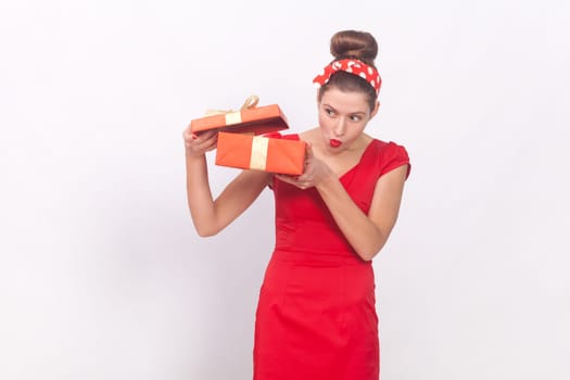 Portrait of curious attractive woman wearing red dress and head band holding present box, looking inside with interest, having gift on her birthday. Indoor studio shot isolated on gray background.