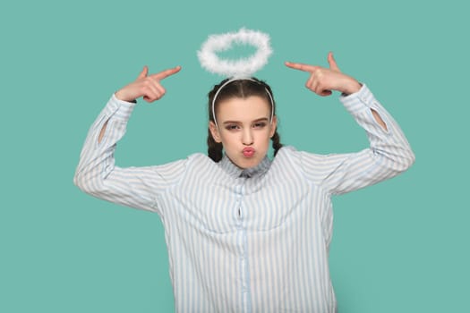 Portrait of funny positive teenager girl with braids wearing striped shirt standing pointing at nimb over her head, looking at camera with pout lips. Indoor studio shot isolated on green background.