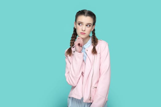 Portrait of pensive thoughtful teenager girl with braids wearing pink jacket standing holding her chin, looking away, planning her future. Indoor studio shot isolated on green background.