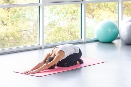 Portrait of flexible woman doing morning yoga after waking up, practices child pose in balasana on pink mat, resting exercise, workout. Indoor shot with window on background.