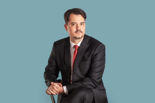 Portrait of handsome serious strict man with mustache sitting on chair, looking at camera with bossy expression, wearing black suit with red tie. Indoor studio shot isolated on light blue background.