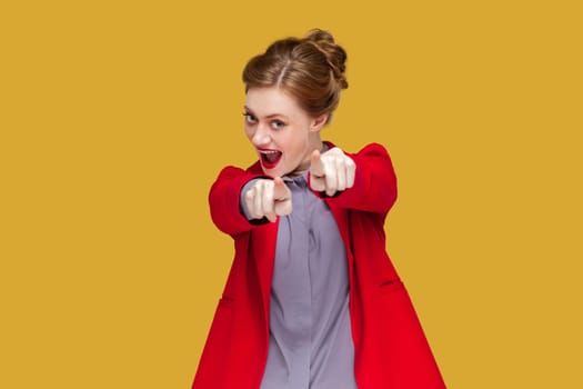 Portrait of excited flirting woman with red lips standing pointing at you, looking at camera with positive expression, wearing red jacket. Indoor studio shot isolated on yellow background.
