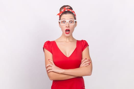 Portrait of shocked surprised woman wearing red dress, glasses and head band standing with crossed arms, looking at camera with big eyes and open mouth. Indoor studio shot isolated on gray background.