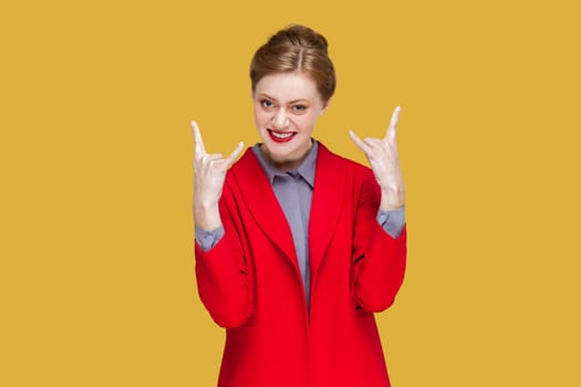 Portrait of excited crazy woman with red lips standing showing rock and roll gesture, having fun on festival, wearing red jacket. Indoor studio shot isolated on yellow background.