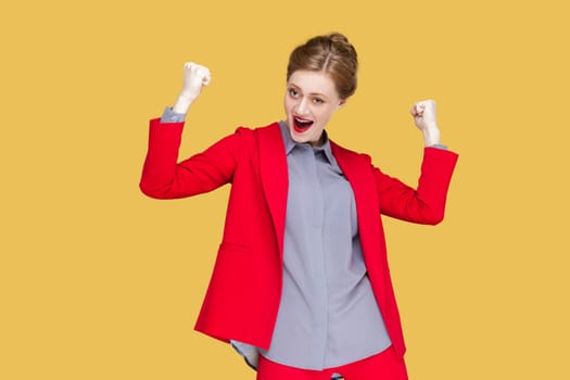 Portrait of extremely happy woman with red lips standing raised her clenched fists, screaming hurray, celebrating her victory, wearing red jacket. Indoor studio shot isolated on yellow background.