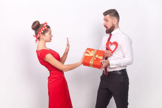 Portrait of shocked sad woman showing stop gesture to man with present box and red heart in hands, female saying no, refuse a gift. Indoor studio shot isolated on gray background.