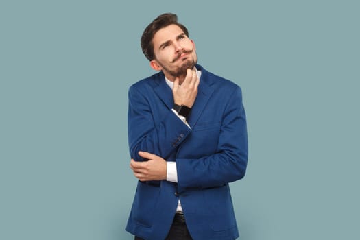 Portrait of thoughtful serious businessman with mustache standing and thinking about his work, holding his chin, wearing official style suit. Indoor studio shot isolated on light blue background.
