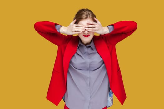 Portrait of shocked woman with red lips standing covering her eyes with palm, do not want to see something scary, wearing red jacket. Indoor studio shot isolated on yellow background.