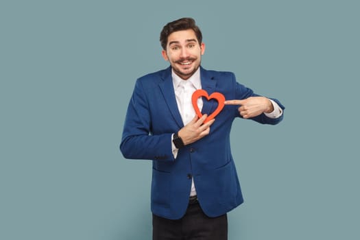 Portrait of funny positive optimistic man with mustache standing pointing at heart figure, looking at camera, wearing white shirt and jacket. Indoor studio shot isolated on light blue background.