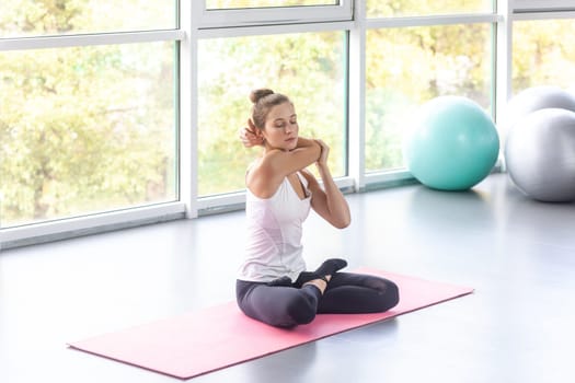Portrait of attractive young woman warming up her hands, training and stretching arm before workout, sitting on yoga mat, doing stretch exercise. Indoor shot with window on background.