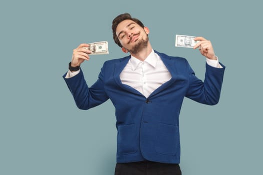 Portrait of happy smiling delighted man with mustache standing with dollar banknotes and looking at camera with pride, wearing official style suit. Indoor studio shot isolated on light blue background