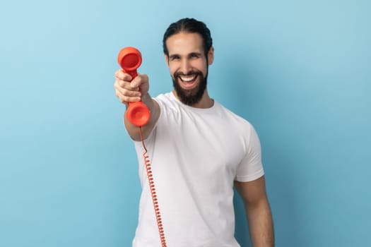 Portrait of handsome happy man with beard wearing white T-shirt holding and showing retro phone handset to camera, asking to answer phone. Indoor studio shot isolated on blue background.